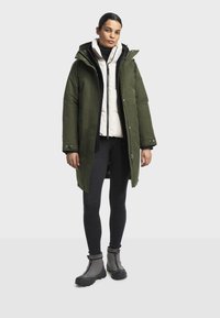 Parka imperméable vert olive avec fermeture éclair à l'avant, grande capuche et poches latérales, superposée à une doudoune blanche. Portée avec des leggings noirs et des bottes grises.