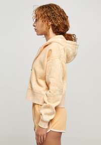 Felpa corta color pesca con un motivo testurizzato e macchiato, dotata di cappuccio con lacci e polsini a costine. Abbinata a pantaloni corti coordinati.
