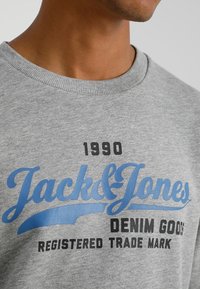 Acercamiento a una sudadera gris con el texto azul y negro "Jack & Jones Denim Goods Marca Registrada 1990" en el pecho.