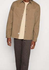 Koszula na guziki w kolorze beżowym z dwoma przednimi kieszeniami, wykonana z wytrzymałej tkaniny. Noszona na jasnobeżowej koszulce i zestawiona z ciemnymi spodniami.