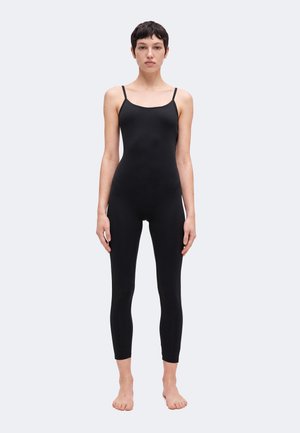 Kvinde stående barfodet iført en tætsiddende sort ærmeløs jumpsuit med tynde skulderstropper mod en ensfarvet lys baggrund.