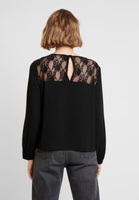 Blusa nera con schiena in pizzo e dettaglio a chiave, maniche lunghe e vestibilità rilassata. Il pizzo presenta un motivo floreale.