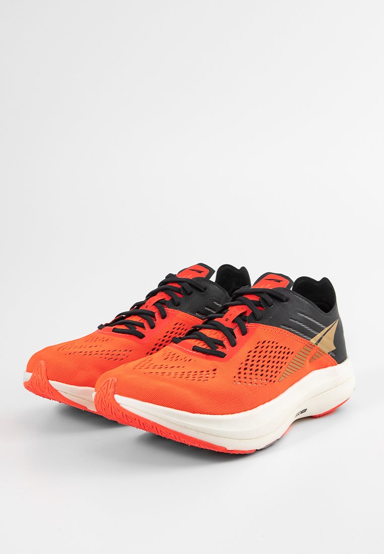 Un par de zapatillas de running naranjas y negras con suelas blancas, cordones negros y un logo dorado a los lados, exhibidas sobre una superficie blanca lisa.