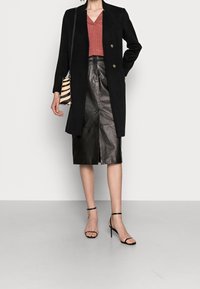 Cappotto in lana nera con bottoni doppi, abbinato a un top in jersey bordeaux con scollo a V e una gonna a matita in pelle nera. Accessoriato con una borsa a righe.