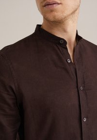 Cămașăbutton-up de culoare maro închis, cu guler mandarin, având un material texturat și detalii subtile de cusătură. Butoanele sunt de nuanță închisă.