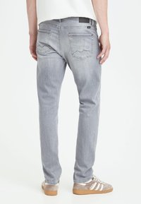 Person iført lysegrå jeans og beige Adidas sneakers, der står med ryggen mod en hvid baggrund.