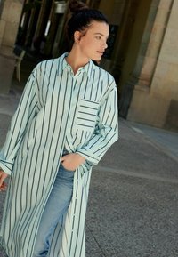 Stribet lang skjorte i lyseblå med lodrette sorte striber, med en brystlomme og knappelukning, parret med denimjeans.