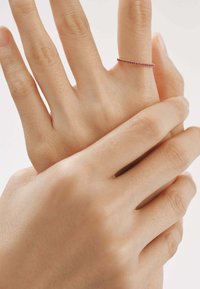 Anillo de oro delicado con pequeñas piedras preciosas rojas, llevado en un dedo. Las manos tienen una textura de piel suave, mostrando el diseño minimalista del anillo.