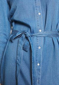 Denim-mekko vaaleansinisellä sävyllä, napitettuna edestä, itsekiinnitettävällä vyötäröhihnalla ja pleat-tyylisellä yksityiskohdalla lisäämään rakennetta.