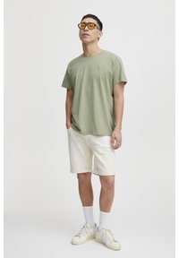 Grønn kortermet t-skjorte med rund hals, beige shorts med oppbrett, hvite joggesko og solbriller med gult skjær. Glatt stofftekstur.