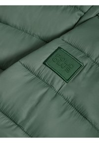 Grön puffjacka i tyg med quiltade sektioner. Har en rektangulär grön broderad etikett med texten "POLO CLUB". Slät struktur.