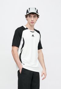 Męski strój sportowy składający się z białej koszulki z krótkim rękawem z czarnymi akcentami i fakturą, w połączeniu z czarnymi spodniami oraz czarno-szarym daszkiem.