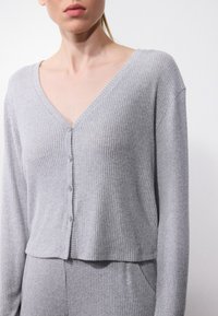 Cardigan gri, cu pliuri, având un decolteu în V, mâneci lungi și închidere cu butoane. Textură moale cu un design scurt, care completează pantaloni asortați.