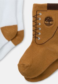 Une paire de chaussettes de type crew, l'une blanche avec un orteil et un talon marron, l'autre marron avec un motif de logo de botte et d'arbre sur la cheville.