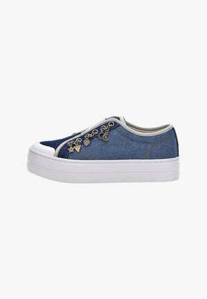 Matala denim tennarit valkoisella platolla, beige reunus ja koristeelliset metalliset riipukset sivuletsissä.