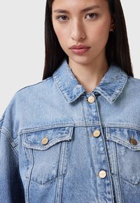 Chaqueta de mezclilla azul claro con cuello, botones frontales y dos bolsillos en el pecho. La textura desgastada añade detalle a la tela.