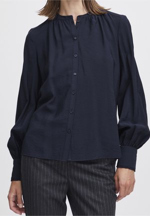 Vrouw met een navyblauwe blouse met knopen en lange pofmouwen en zwarte broek met fijne strepen, getoond van hals tot heupen.