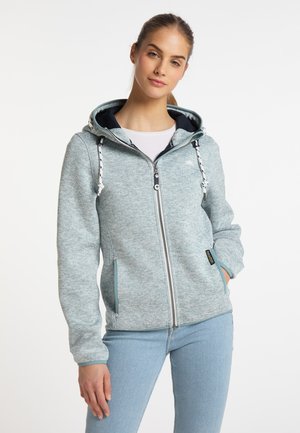 Světle šedá mikina na zip s texturovanou tkaninou, s kapucí, šňůrkami a bočními kapsami. Kombinována se světle modrými džíny.
