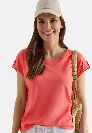 Smiling woman with long brown hair wearing a coral short-sleeve shirt, beige cap reading "OPTIMISM," and carrying a woven shoulder bag.