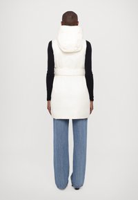 White padded vest with a hood, belted waist, and smooth texture. Paired with light blue wide-leg jeans. Minimalist design and no visible embellishments.