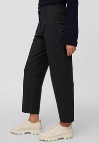 Persoon in zwarte, op maat gemaakte broek en crèmekleurige sneakers met een dikke zool, met een mouw van een marineblauwe trui zichtbaar tegen een neutrale achtergrond.