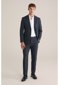 Tumši zils uzvalks no teksturētas auduma, ar vienrindu žaketu ar ieliektiem atlokiem un piegrieztām biksēm, kopā ar baltu kreklu.