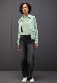 Kobieta z kręconymi włosami, ubrana w miętową zieloną kurtkę i sweter, szerokie czarne dżinsy oraz białe sneakersy, stojąca przy ciemnoszarym murze.