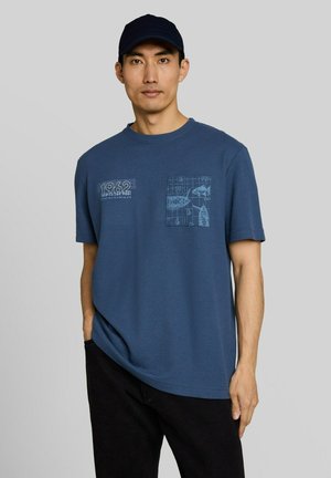 Man draagt een blauw grafisch T-shirt en zwarte broek met één hand in de zak, draagt een zwarte pet, staat tegen een effen grijze achtergrond.