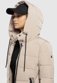 Hellbeige Pufferjacke mit Kapuze, aus strukturiertem Stoff, ausgeprägter Naht und schwarzen Kordeln. Das Model trägt eine schwarze Mütze.