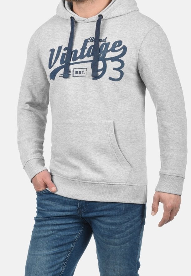 Homme portant un sweat à capuche gris clair avec des cordons bleu marine et le texte "Vintage 93", associé à un jean bleu, debout avec une main dans la poche.