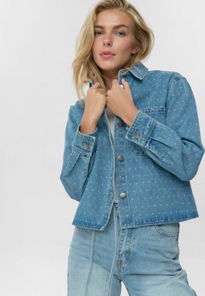 Light blue denim jacket with a button-up front, two pockets, and a slightly cropped fit. Fabric features a subtle polka dot pattern.