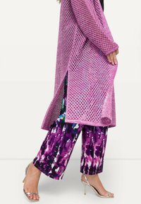 Cárdigan largo de punto abierto en rosa brillante, superpuesto a unos pantalones con estampado colorido en púrpura y blanco, complementado con sandalias de tacón alto plateadas.