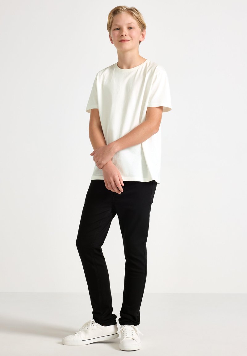 Bílá bavlněná tričko s krátkým rukávem, spárované s černými slim-fit kalhotami a bílými teniskami. Čistý design, bez viditelných vzorů nebo akcentů.