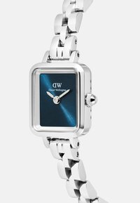 Sidabrinis stačiakampis laikrodis su mėlynu metaliniu ciferblatu, turintis sidabrines rodykles ir tekstūruotą grandinėlę. Prekės ženklas „Daniel Wellington“ matomas.