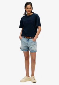 Marine crew-neck t-shirt med broderet logo, parret med lyseblå denimshorts med frynset kant og beige sneakers.