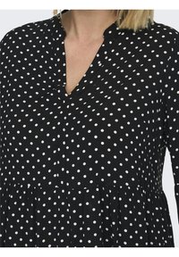 Schwarzes Kleid mit weißen Punkten, mit einem V-Ausschnitt, Knopfverschluss und geraffter Taille. Das Material scheint weich und leicht zu sein.