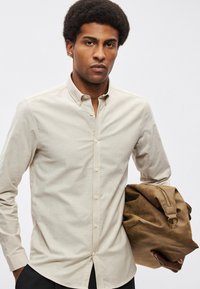 Μπεζ πουκάμισο με κουμπιά, μυτερό γιακά, μακριά μανίκια και στενή εφαρμογή. Κρατάει καφέ δερμάτινη τσάντα με φερμουάρ και ιμάντα ώμου.