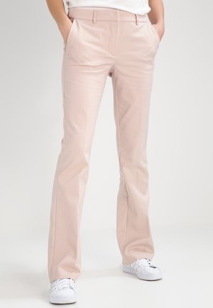 Personne portant un pantalon droit rose clair et des baskets blanches, les mains décontractées dans les poches, debout sur un fond blanc uni.