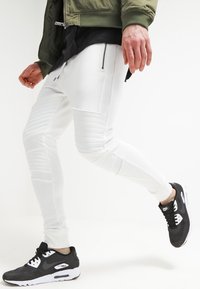 Bílé tapered joggingové kalhoty s vroubkovanými detaily, spárované s černými teniskami od Nike. Kalhoty mají zipové kapsy a hladký povrch.