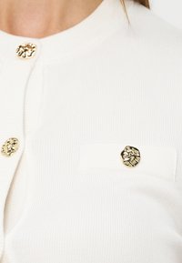 Gros plan sur un cardigan blanc à côtes avec des boutons dorés texturés et irréguliers, dont un sur une poche poitrine.