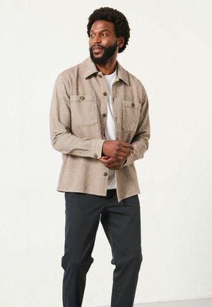 Brown checkered shirt jacket with two chest pockets, button front, long sleeves, and a smooth texture, paired with dark pants.