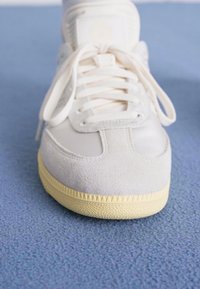 White sneaker featuring a suede and mesh upper, round toe, flat laces, and a textured rubber sole with a yellow accent.