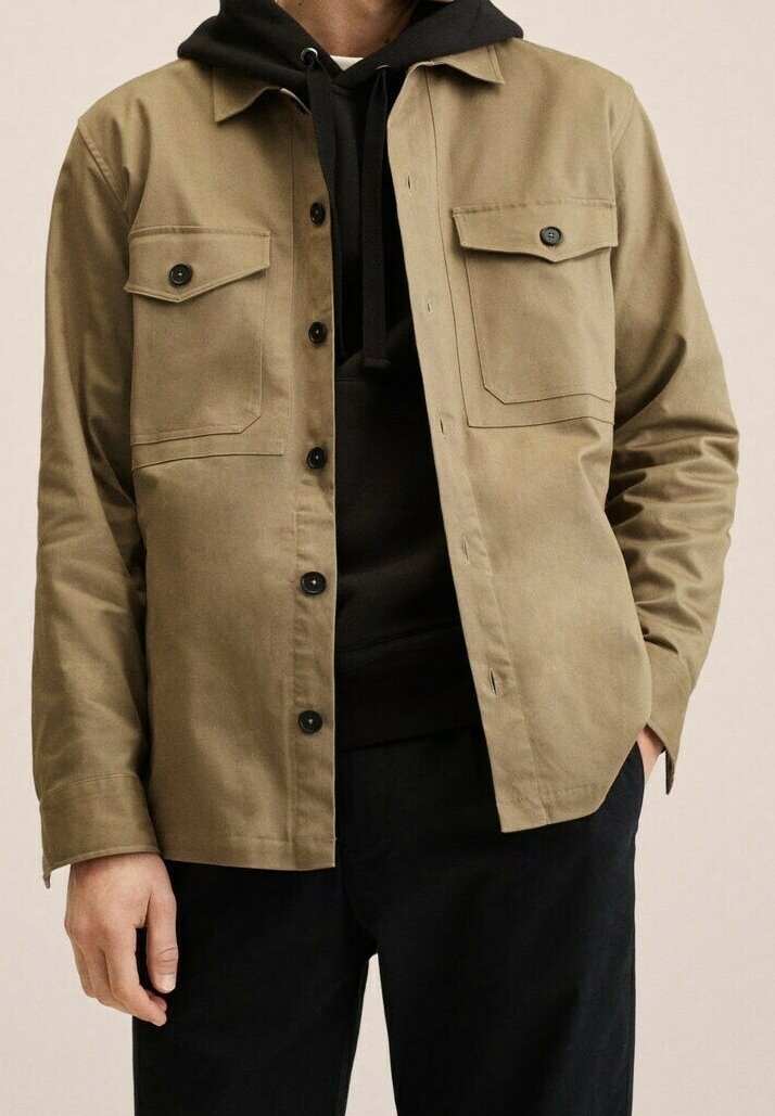 Homme portant une veste beige à boutons par-dessus un sweat à capuche noir, une main dans la poche et un pantalon noir.
