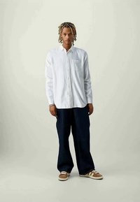 Jovem com dreads loiros usa uma camisa branca de botão, calças pretas largas e ténis castanhos, de pé contra um fundo liso.