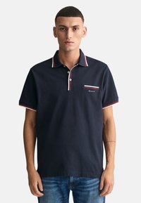 Camisa polo azul marino de algodón con una tapeta de dos botones y cuello a rayas. Presenta un pequeño logo de GANT en el bolsillo del pecho.