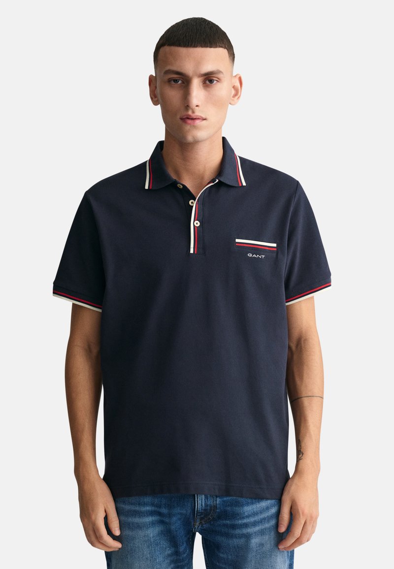 Camisa polo azul marino de algodón con una tapeta de dos botones y cuello a rayas. Presenta un pequeño logo de GANT en el bolsillo del pecho.