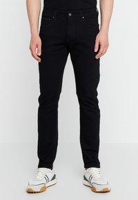 Jeans in denim nero dal taglio slim, design a cinque tasche, chiusura con bottone e cuciture a contrasto. Indossati con scarpe da ginnastica bianche e verdi.