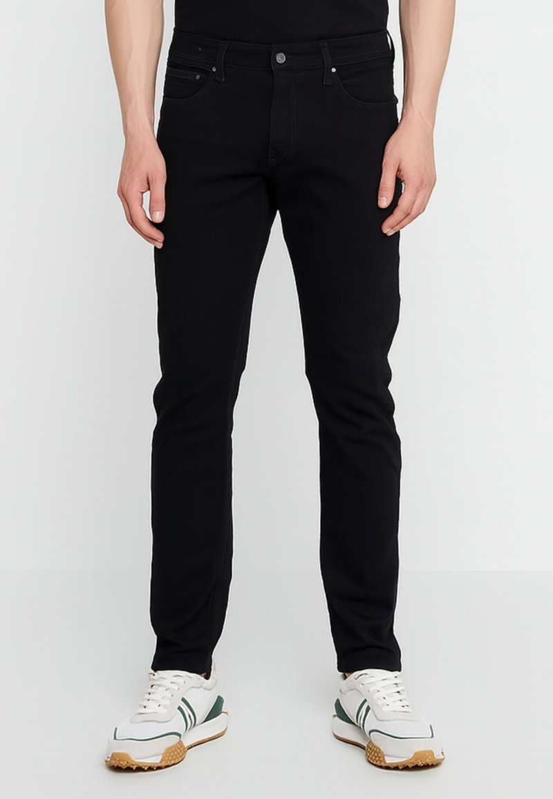 Jeans in denim nero dal taglio slim, design a cinque tasche, chiusura con bottone e cuciture a contrasto. Indossati con scarpe da ginnastica bianche e verdi.