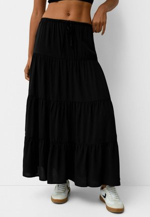 Jupe longue noire à volants avec taille élastique et cordon de serrage. Confectionnée en tissu doux avec une coupe fluide. Portée avec des baskets blanches.