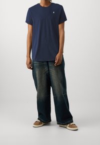 Granatowy bawełniany t-shirt z krótkimi rękawami i okrągłym dekoltem, zestawiony z luźnymi, wyblakłymi niebieskimi jeansami i sneakersami z brązowymi akcentami.